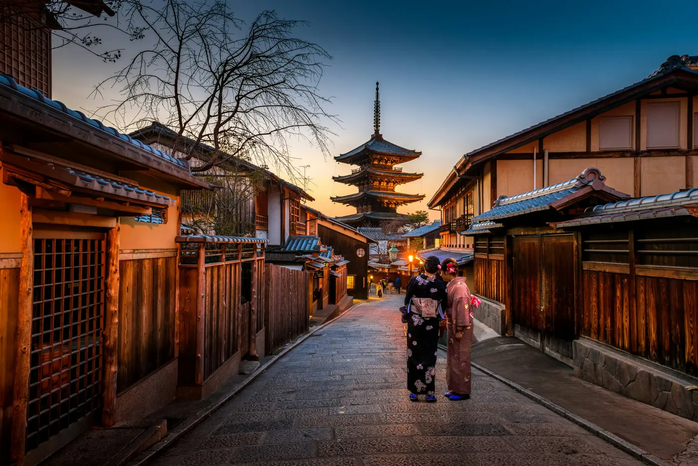 Two people in kimono standing on a narrow street in Kyoto, with a pagoda behind them.